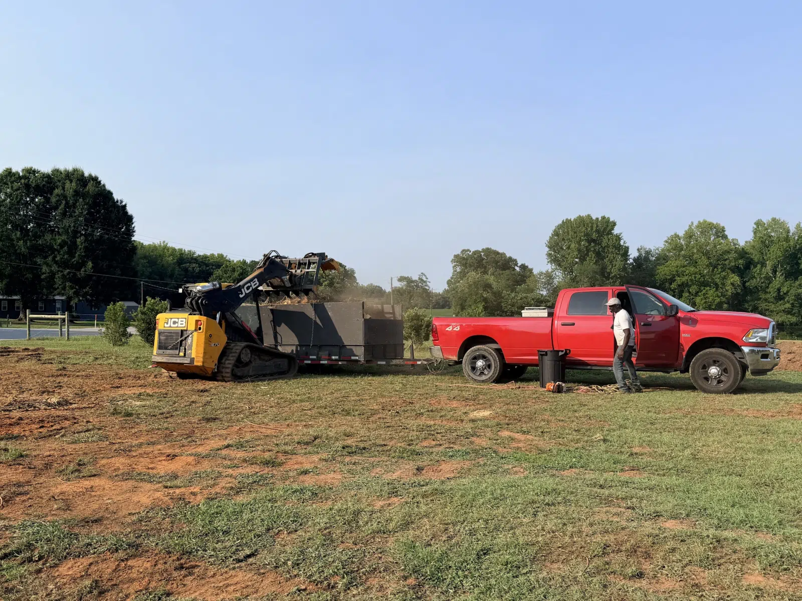Skid steer loading debris into a trailer beside a red truck — Your Way Lawn and Tree in Greensboro